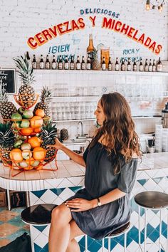 a woman sitting at a table in front of a counter filled with oranges and pineapples