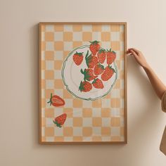 a woman holding up a painting with strawberries on it in front of a checkered wall