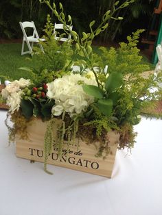 a wooden box filled with flowers and greenery on top of a white table cloth