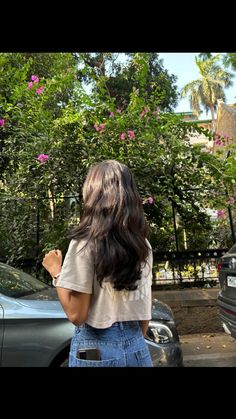 a woman standing in front of a car with her back to the camera and looking at pink flowers