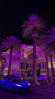 a blue sports car driving past palm trees in front of a large building at night