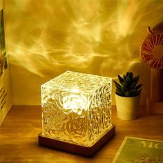 an illuminated cube sitting on top of a wooden table next to a book and potted plant