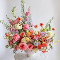 A flower arrangement in a glazed white compote vase in front of a white backdrop on a marble table. It's filled with pink, yellow, peach, orange, blue, purple, and white-colored flowers, including rose, ranunculus,, piccolini daisy, tulip, oncidium orchid, chamomile, snapdragon, and delphinium. The vibes are very Bridgerton. Spring Wild Flower Wedding Bouquet, Eucalyptus Garland With Bright Flowers, Wildflower Bridesmaid Flowers, Flowers For June Wedding Bridal Bouquets, Wedding Flower Inspirtion, Boho Wild Flower Bouquet, What Flowers Are Popular For A May Wedding, Every Flower Wedding Bouquet, Bright Summer Floral Arrangements