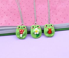 three green frog necklaces with flowers on them sitting on a pink tablecloth next to a book