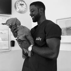 a man holding a baby in his arms while standing next to a wall with clocks on it