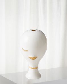 a white vase with gold details on the top and bottom, sitting on a table in front of a curtain