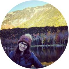 a woman wearing glasses and a knitted hat stands in front of a mountain lake