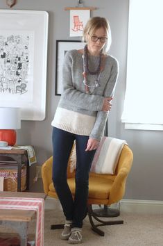 a woman standing in a living room next to a yellow chair and table with pictures on the wall