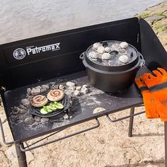 an outdoor grill with food cooking on it next to a body of water and two chairs