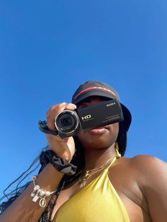 a woman in a yellow top holding a camera up to her face with the sky behind her