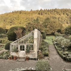 an old greenhouse in the middle of a garden surrounded by trees and shrubs with mountains in the background