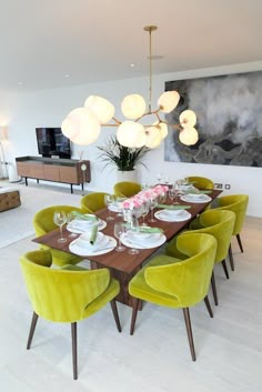 a dining room table with green chairs and white plates on it, in front of a large painting