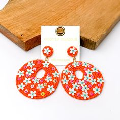 Orange beaded circular drop earrings with pink and blue designs. Pictured on a white background with a wood piece at the top. Trendy Round Beaded Earrings, Trendy Circle Earrings For Spring, Trendy Circular Beaded Earrings, Multicolor Beaded Flower Earrings For Spring, Spring Multicolor Beaded Flower Earrings, Spring Beach Jewelry With Round Shape, Trendy Pink Round Beaded Earrings, Trendy Circular Earrings With Colorful Beads, Multicolor Round Earrings For Spring