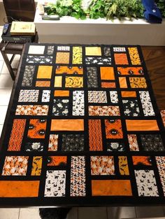 an orange and black patchwork quilt on a table with potted plants in the background