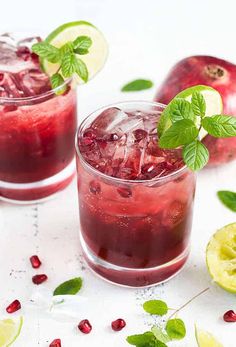 two glasses filled with ice and garnish next to sliced lemons, pomegranate and mint