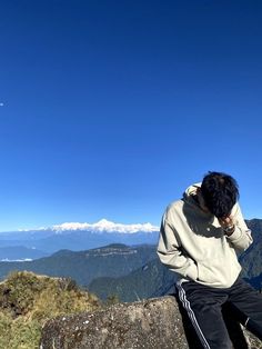 a man sitting on top of a large rock next to a lush green hillside under a blue sky