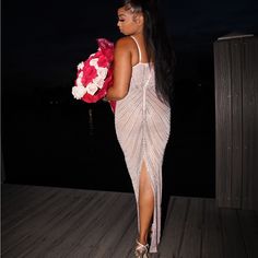 a woman in a long dress holding flowers on a dock at night time with her back turned to the camera