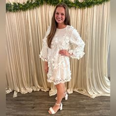 a woman standing in front of a backdrop wearing a white dress with flowers on it