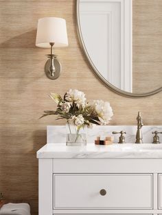 a white bathroom vanity with flowers in a vase on the counter and a round mirror above it