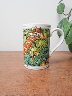 a coffee cup with a giraffe painted on it sitting next to a potted plant