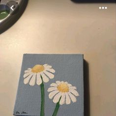 two white daisies painted on a blue square with yellow centers sitting on a table