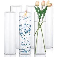 three clear vases with flowers in them on a white surface, one has a lit candle and the other is filled with water