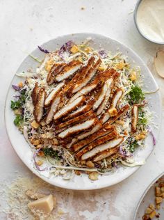 a white plate topped with chicken and coleslaw next to two bowls of dressing