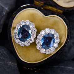 This flattering pair of earrings each feature one (1) oval cut natural sapphire set into a four-prong setting. The sapphires are each bordered with twelve (12) semi-bezel/ bead set round brilliant cut diamond accents. The diamonds are bordered with milgrain edging. The earrings measure 12.4mm by 10.5mm and are finished with friction backs. Luxury Oval Sapphire Diamond Earrings, Oval Diamond Cluster Earrings For Formal Events, Luxury Oval Diamond Earrings For Anniversary, Elegant Oval Cluster Earrings With Brilliant Cut, Classic Oval Cluster Earrings For Formal Occasions, Luxury Oval Diamond Earrings With Gemstones, Classic Blue Oval Diamond Earrings, Luxury Oval Cluster Earrings For Formal Occasions, Oval Halo Design Cluster Earrings For Formal Occasions