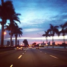 the sun is setting behind palm trees on the road in front of some tall buildings