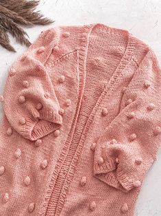 a pink knitted sweater with buttons and a feather on the floor next to it