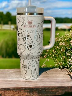 a white travel mug with drawings on it sitting on a wooden table in front of some flowers