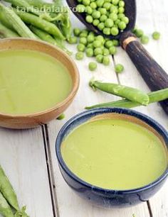 two bowls filled with green pea soup next to peas and spoons on a white wooden table