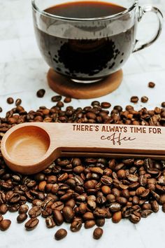 A coffee scoop made from wood with the text: there's always time for coffee, engraved on it. The spoon is sitting on a bed of coffee beans.