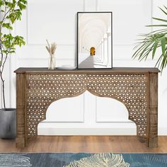 an ornate wooden console table in front of a white wall and potted plant next to it