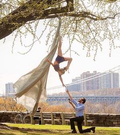 two people doing aerial acrobatic tricks in front of a tree with the brooklyn bridge in the background