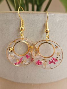 two gold hoop earrings with pink flowers on them sitting on a white marble surface next to a potted plant