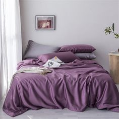 a bed covered in purple sheets and pillows next to a potted plant on a table