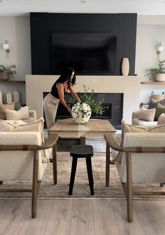 a woman arranging flowers in a vase on a table with two chairs and a fireplace