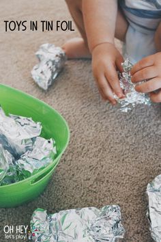 a toddler playing with tin foil on the floor next to a green bowl filled with silver foil