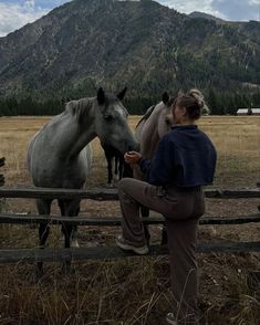 a woman standing next to two horses on top of a dry grass covered field with mountains in the background