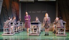 children sit in chairs on stage with teacher standing next to them and writing on the wall behind them