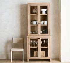 a wooden cabinet with glass doors next to a white chair and vase on the floor