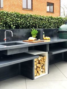 an outdoor kitchen with black counter tops and wood stacked on the outside, in front of a brick building