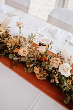 an arrangement of flowers and greenery is arranged on a long table with white linens