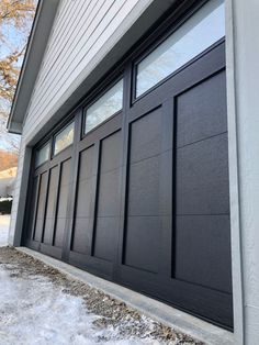 a black garage door on the side of a house with snow around it and trees in the background