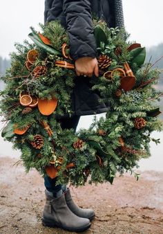 a person holding a wreath with pine cones and oranges on it in front of a body of water