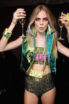 a woman in green and gold outfit holding two wine glasses up to her face with both hands