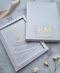 two white and gold wedding cards in a box on a marble surface with flowers next to it