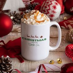 a white coffee mug filled with whipped cream and topped with caramel sauce, sitting on a table surrounded by christmas decorations