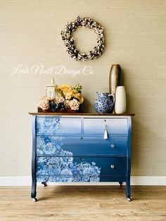 a blue and white dresser with flowers on top next to a wreath hanging on the wall